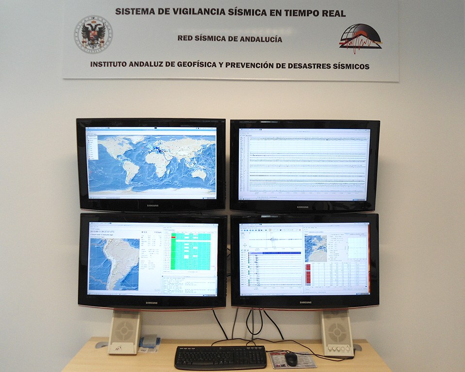 4 monitores bajo un cartel indicando que se trata de equipos de vigilancia sísmica en tiempo real